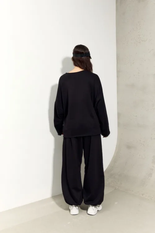 Person with their back to the camera, wearing an oversized black top and wide black pants, white sneakers, and a black headband, standing against a white wall and gray concrete surface. Alternate: Back view of a person in loose black outfit and white sneakers in a minimal studio setting.