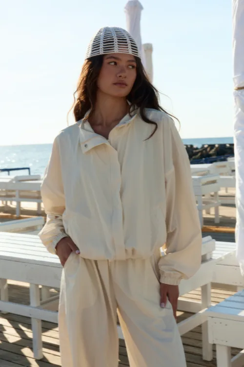 Woman in a light beige jumpsuit standing on a wooden pier by the sea, wearing a white woven hat and looking to the side.