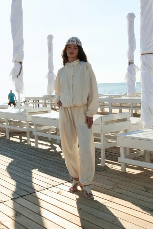 Woman in a light beige jogger set and netted cap stands on a sunlit wooden pier by the sea, with wrapped white posts around her