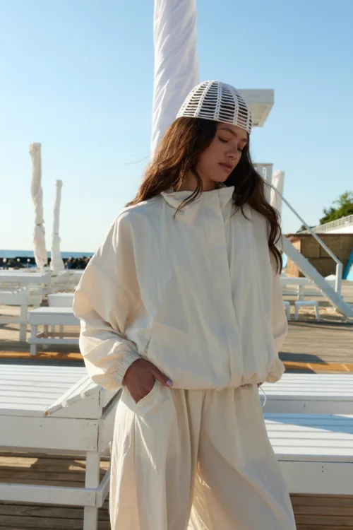 Woman in a light beige oversized jacket and white woven cap stands on a sunlit boardwalk with white outdoor furniture nearby.