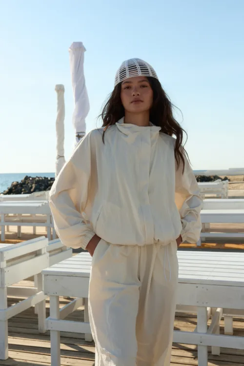 Woman standing outdoors in a white jumpsuit and white netted cap on a seaside boardwalk, with blue sky and ocean in the background.