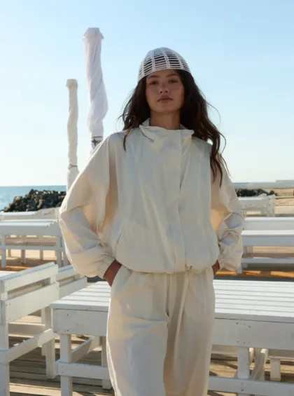 Woman standing outdoors in a white jumpsuit and white netted cap on a seaside boardwalk, with blue sky and ocean in the background.