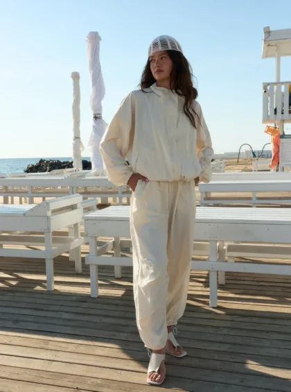 Woman in an off-white hooded jumpsuit and white sandals stands on a wooden beach boardwalk, with a blue sky and ocean in the background.