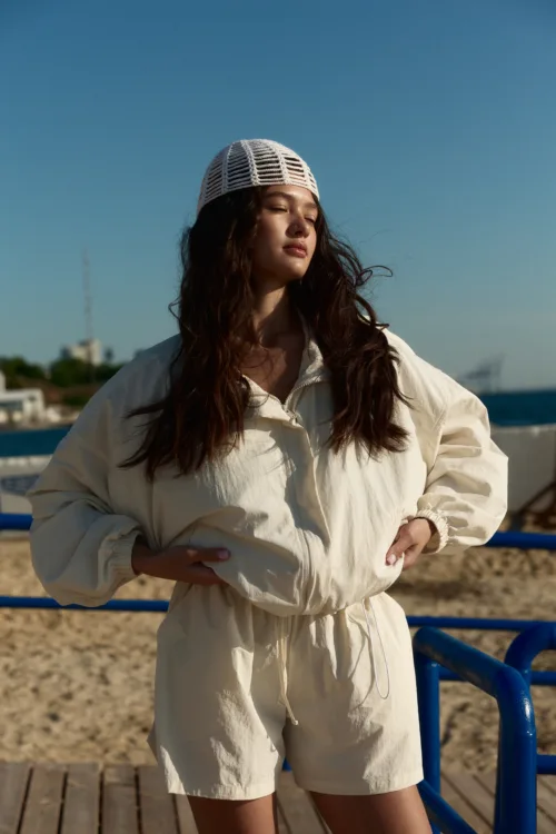 Woman in a cream-colored outfit and white knit cap stands on a wooden boardwalk with the sea in the background, facing the sun.