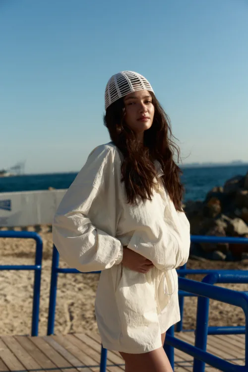 Woman posing at a seaside boardwalk in a white dress and a woven white hat, with the ocean and blue railings in the background.