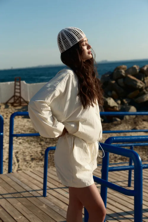 Woman in a white dress and mesh hat stands by a blue railing on a wooden boardwalk near the sea, facing sunset breeze.