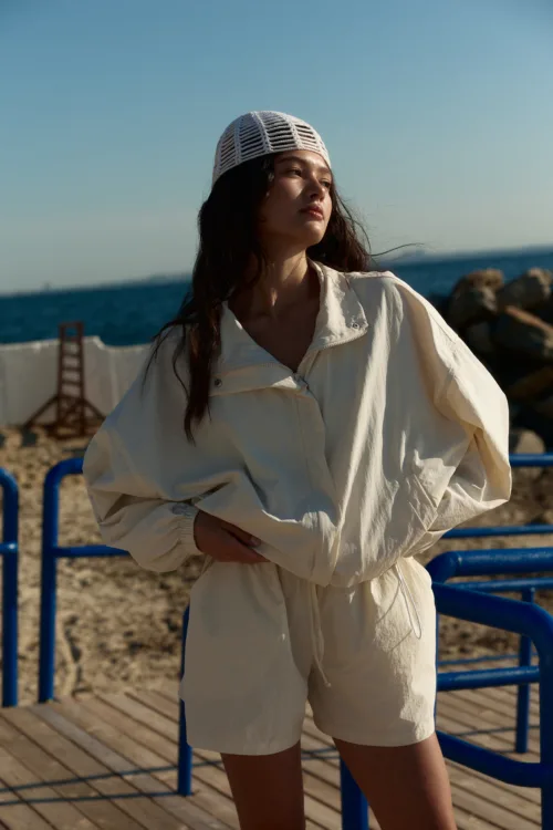 Woman standing on a wooden boardwalk by the sea, wearing a beige jumpsuit and a white knit cap, looking into the distance.