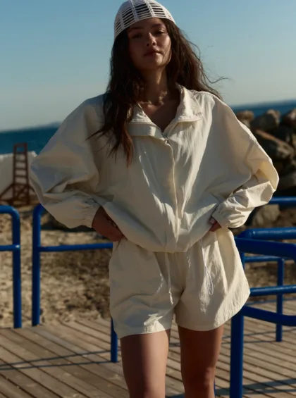 Woman standing on a wooden boardwalk by the ocean, wearing a white jacket and a white knit cap, hands on hips.