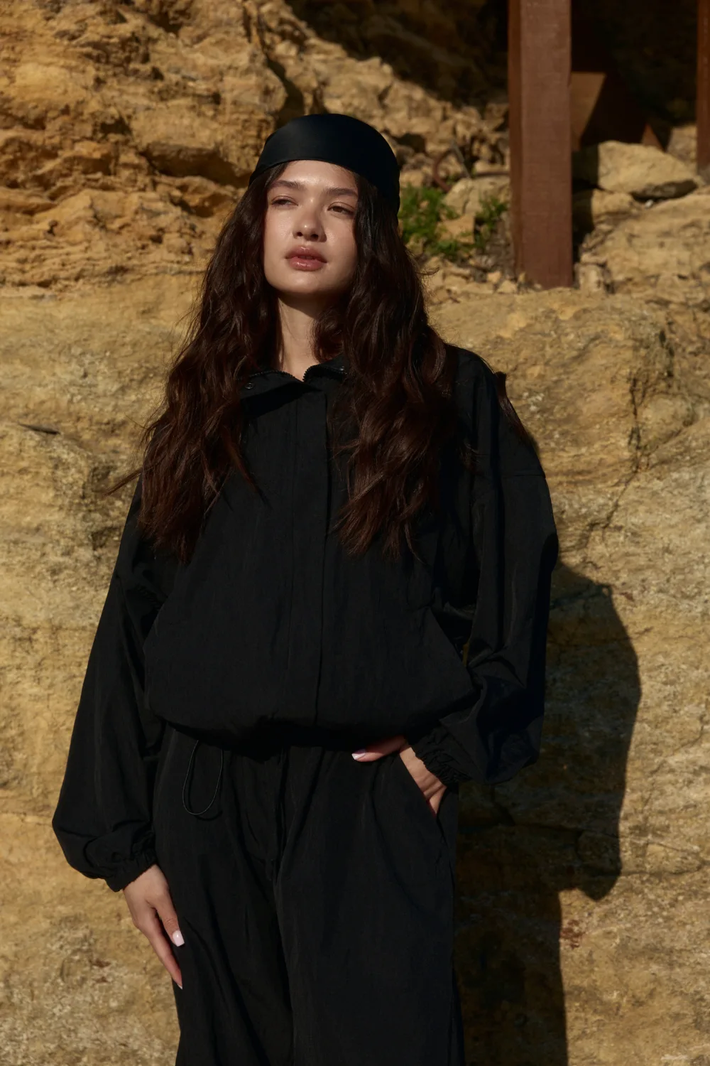 Woman in a black outfit and headscarf standing outdoors against a rocky, sunlit backdrop.