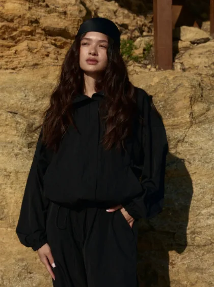 Woman in a black outfit and headscarf standing outdoors against a rocky, sunlit backdrop.