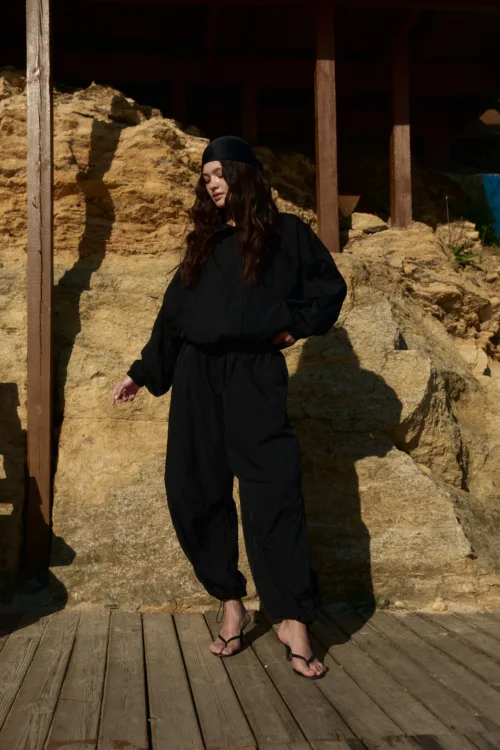 Woman in black outfit and headscarf stands on a wooden deck beside a rocky, sunlit backdrop.
