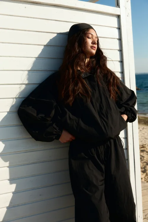 Woman in a black outfit leaning against a white wooden wall by the beach, eyes closed in sunlight.