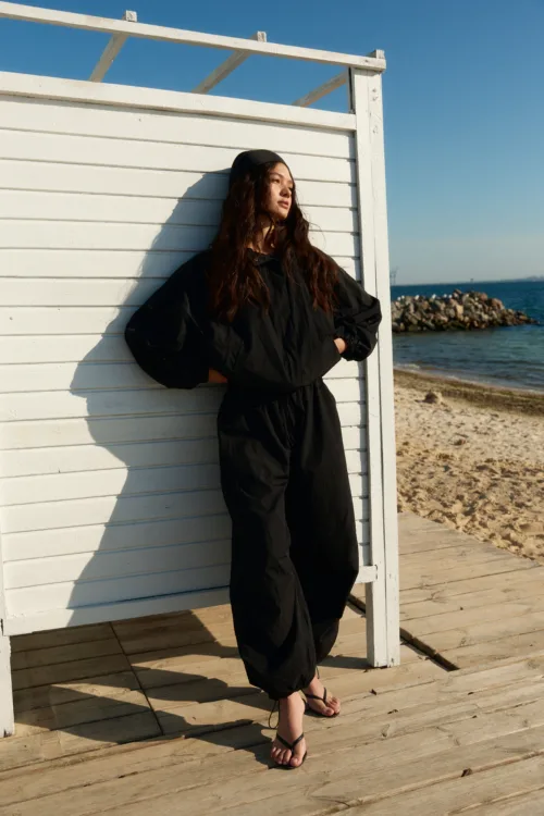 Woman in a black jumpsuit leaning against a white wooden panel wall on a sunny beach, with blue sky and ocean in the background.