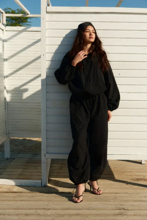 Woman in a black, baggy jumpsuit standing on a wooden deck beside a white slatted fence, wearing strappy heeled sandals in sunlight.