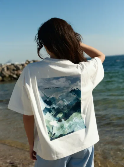 Person facing away from camera at the seaside, wearing a white T-shirt with a blue abstract landscape graphic on the back.