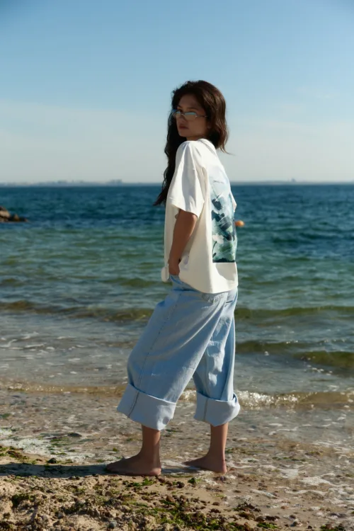 Woman standing barefoot on a sandy beach in a white T‑shirt and light blue jeans, with the ocean in the background.