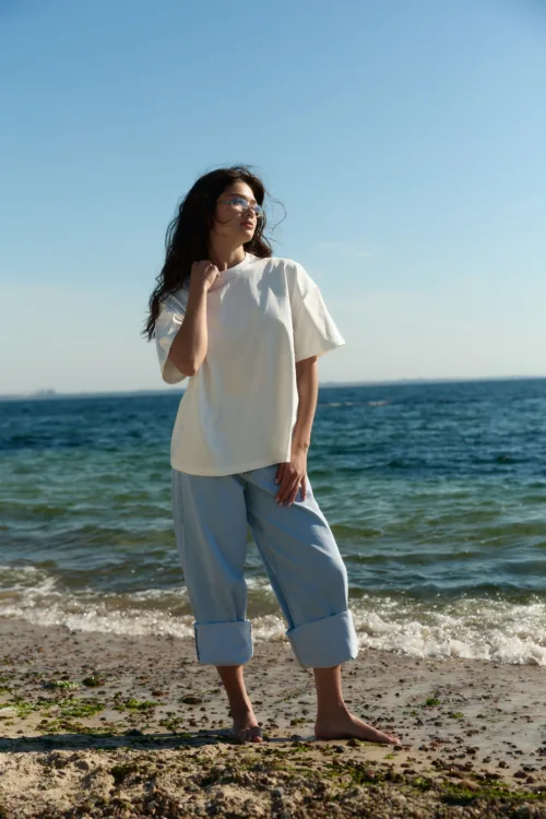 Woman standing barefoot on a sandy beach, looking out toward the ocean, wearing a loose white top and light jeans.
