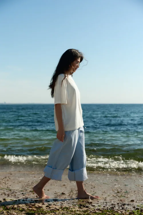 Woman walking barefoot on a rocky beach in a white shirt and light blue rolled-up jeans, with the ocean behind her.