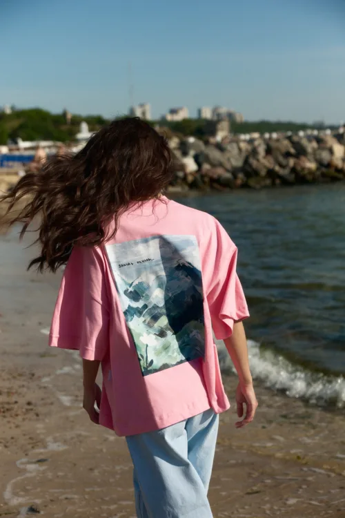 Person with long wavy hair wearing a pink shirt with a graphic on the back, standing on a beach by the water, facing away from the camera.