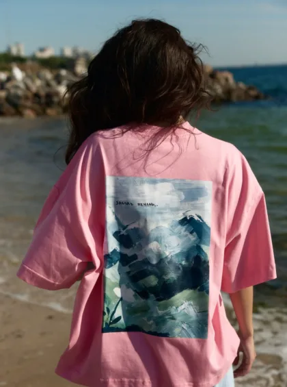 Person seen from behind on a sunny beach, wearing a pink shirt with a blue-green abstract graphic on the back and dark hair blowing in the breeze.