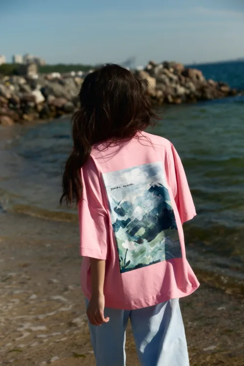 Back view of a person with long dark hair wearing a pink oversized shirt with a graphic on the back, by a rocky shoreline and sea behind them.