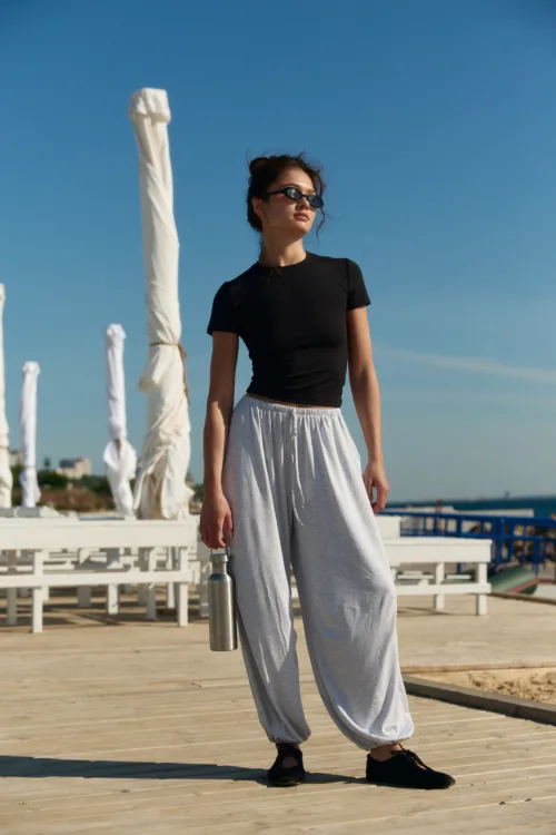 Woman wearing a black t-shirt and gray joggers stands on a sunny beach boardwalk, holding a metal water bottle and wearing sunglasses.