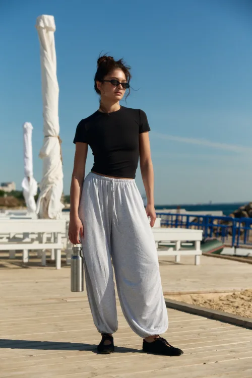 Woman standing on a wooden boardwalk near the beach, wearing a black short-sleeve top, gray wide-leg pants, and sunglasses, holding a metal water bottle.