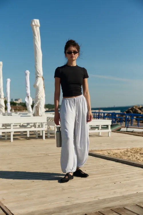 Woman standing on a wooden boardwalk at the beach, wearing a black top, gray sweatpants, black shoes, and sunglasses.