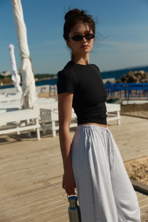 Woman in a black top and gray skirt standing on a wooden boardwalk by the sea, wearing sunglasses and holding a water bottle.