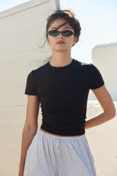 Woman wearing a black short-sleeve top and light gray skirt, sunglasses on, standing outdoors beside a white wall on a sunny day.