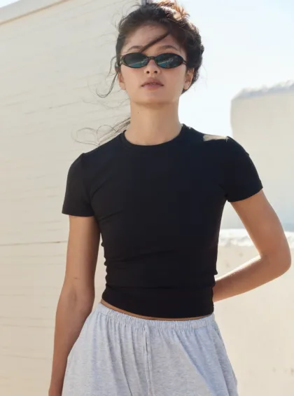 Woman wearing a black short-sleeve top and light gray skirt, sunglasses on, standing outdoors beside a white wall on a sunny day.