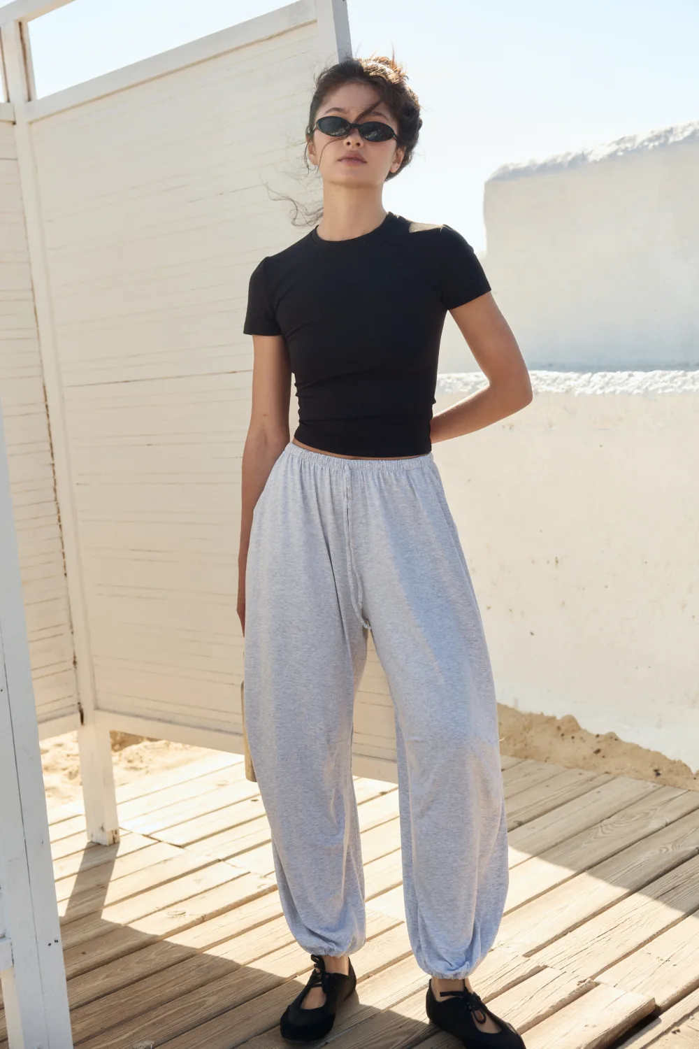 Woman standing outdoors on a sunlit wooden deck, wearing a black short-sleeve top, light gray jogger pants, black shoes, and sunglasses, looking at the camera.
