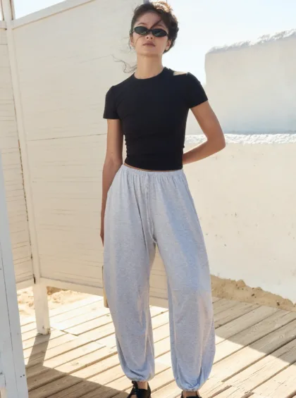 Woman standing outdoors on a sunlit wooden deck, wearing a black short-sleeve top, light gray jogger pants, black shoes, and sunglasses, looking at the camera.