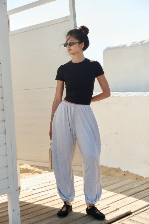 Woman wearing a black cropped top and gray sweatpants stands on a wooden deck beside a white fence, wearing sunglasses on a sunny day.