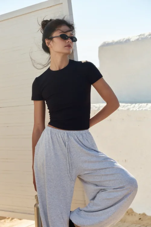Woman posing outdoors in a black fitted t-shirt and light gray sweatpants, wearing dark sunglasses beside a white wall.