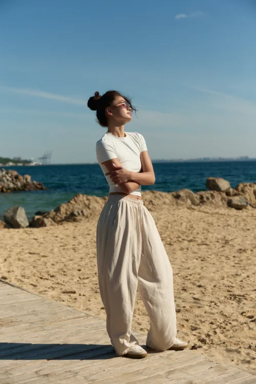 Woman standing on a wooden boardwalk at the beach, wearing a white t-shirt and light beige pants, arms crossed, looking toward the sea with sunglasses on.