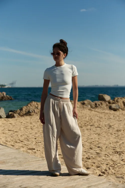 Woman standing on a sandy beach near the water, wearing a white short-sleeve top, beige wide-leg pants, and sunglasses with her hair in a bun, rocks and calm sea in the background.