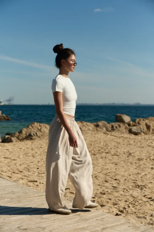 Woman walking along a sandy beach on a wooden boardwalk, wearing a white top, beige pants, sunglasses, and her hair in a bun (side profile).] ,