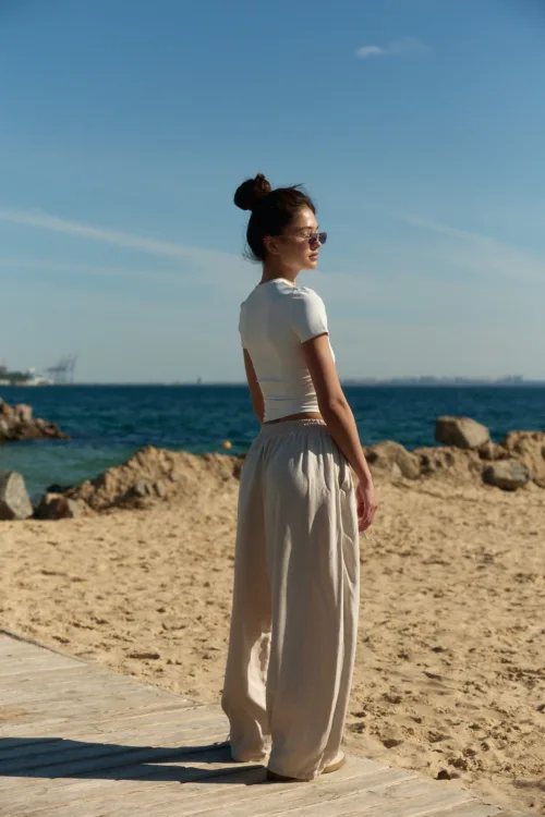 Woman standing on a sandy beach by the ocean, wearing a white short-sleeve top, beige wide-leg pants, and sunglasses, facing right.