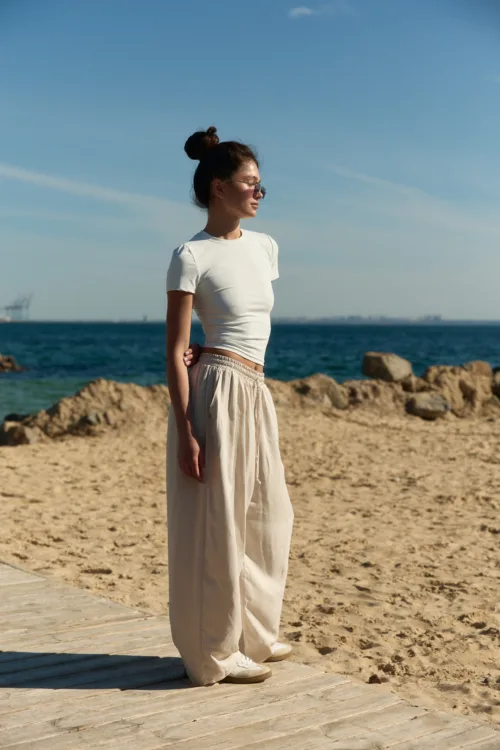 Woman standing on a sandy beach beside a wooden boardwalk, facing right, wearing a white top and beige wide-leg pants with sunglasses; sea and rocks in the background.