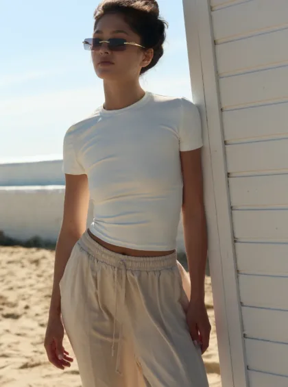 Person wearing a white fitted top and light drawstring pants stands beside a white beach shack on a sunny day. But the alt text should describe the image's meaning, not just appearance.