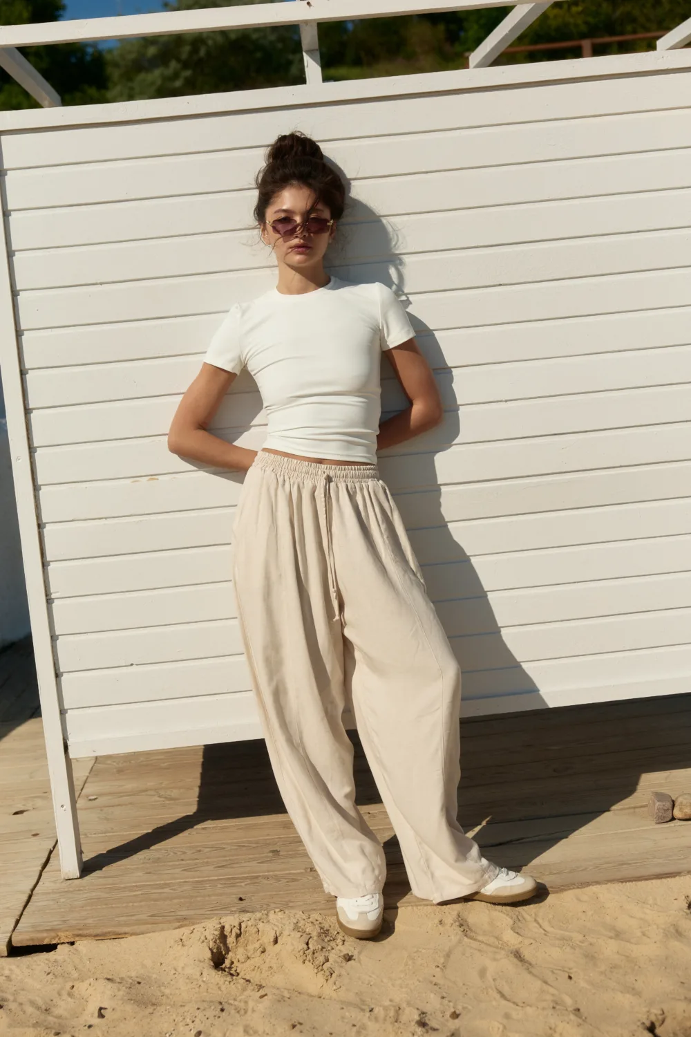 Woman wearing a white top and beige wide-leg pants stands against a white wooden fence on a sunny beach.