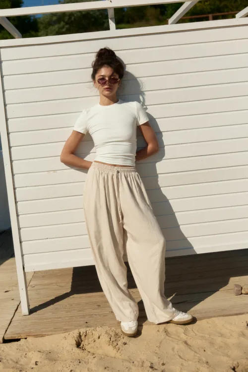 Woman wearing a white top and beige wide-leg pants stands against a white wooden fence on a sunny beach.