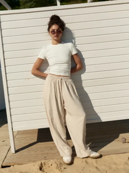 Woman wearing a white top and beige wide-leg pants stands against a white wooden fence on a sunny beach.