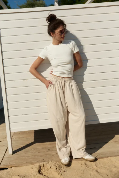 Woman with dark hair in a bun, wearing a white short-sleeve top and beige wide pants, leaning against a white slatted wall at the beach with sunglasses.