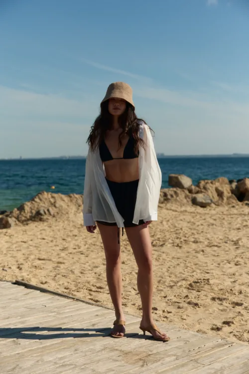 Woman in a black bikini top and white cover-up stands on a wooden boardwalk at the beach, with the sea in the background.