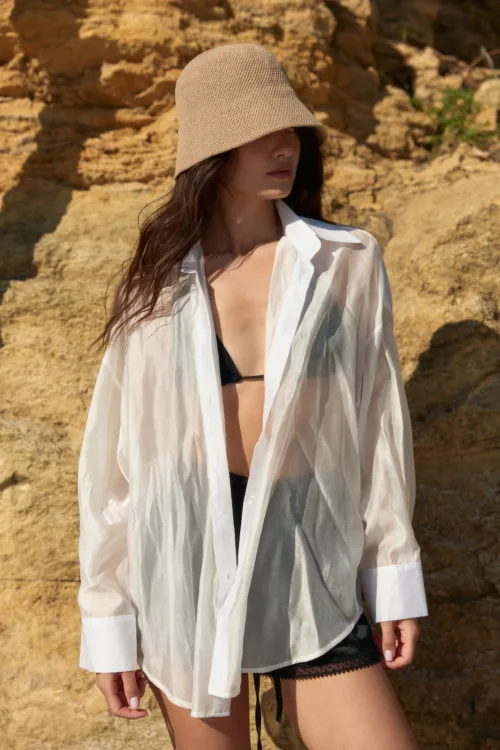 Woman wearing a beige woven bucket hat and a sheer white shirt open at the chest, standing against rocky cliffs; black lingerie shows beneath.