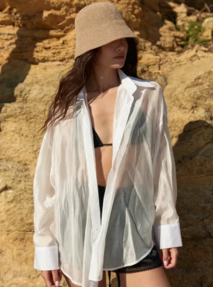 Woman wearing a beige woven bucket hat and a sheer white shirt open at the chest, standing against rocky cliffs; black lingerie shows beneath.