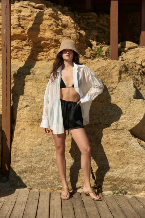 Woman posing on a rocky beach setting in a beige sunhat, white shirt, black bikini top and black shorts with tan sandals.