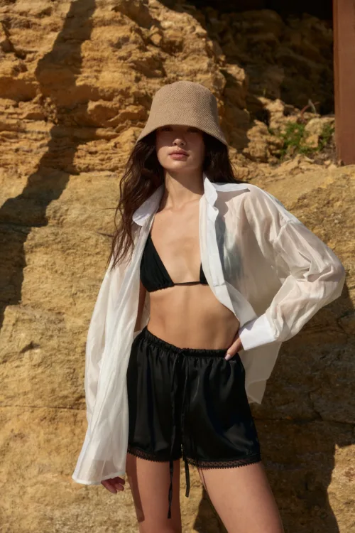 Woman in a black bikini top and sheer white shirt stands against rocky beige cliffs, wearing a beige bucket hat.